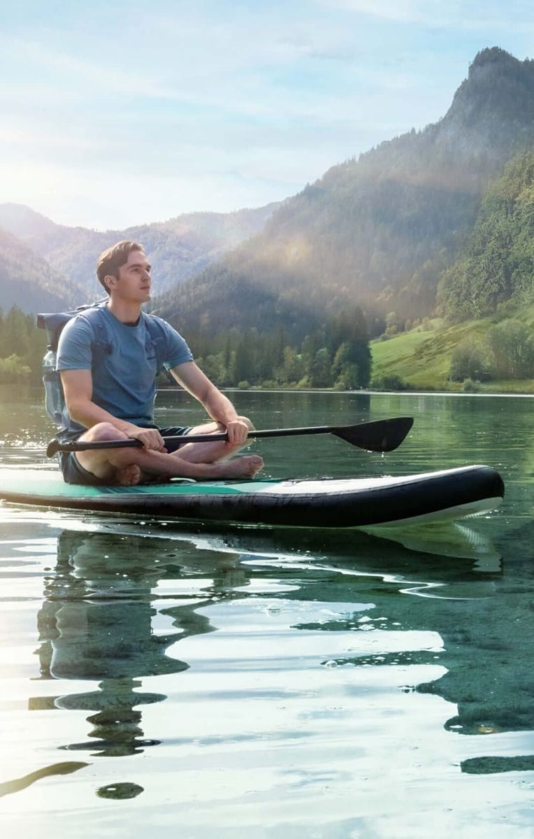 Ein Mann in blauem T-Shirt sitzt im Schneidersitz auf seinem SUP auf einem türkis-blauen See umringt von Bergen und Wäldern. Der Himmel ist blau und die Sonne scheint auf den See.