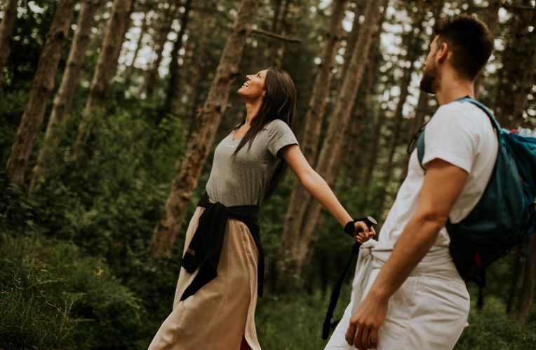Ein Paar genießt einen Spaziergang im Wald – die Frau blickt lächelnd nach oben, während sie die Natur spürt, der Mann trägt einen Rucksack und hält ihre Hand.
