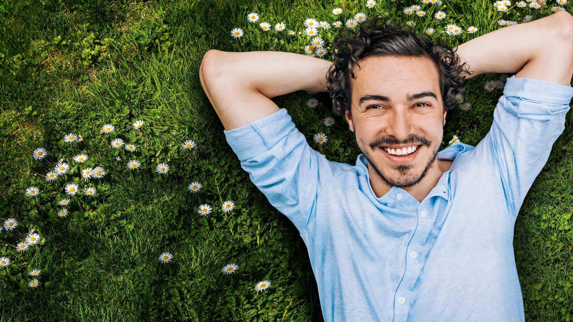 junger Mann mit schwarzen Haaren und blauen Hemd schaut in die Kamera und liegt auf einer grünen Wiese mit Gänseblümchen